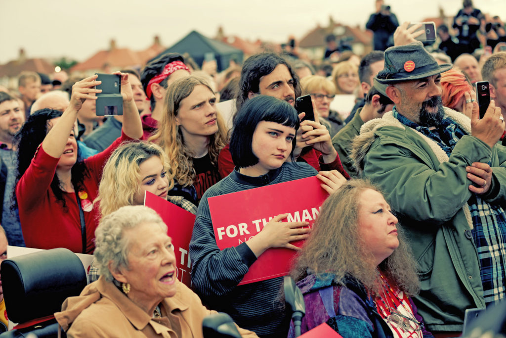 Young woman holding 'For the Many' placard among crowd listening to Jeremy Corbyn