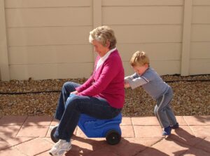 Smiling grandmother in red jumper being pushed by young grandson, , By Catherine Scott - Matti, CC BY-SA 2.0, https://commons.wikimedia.org/w/index.php?curid=12595647